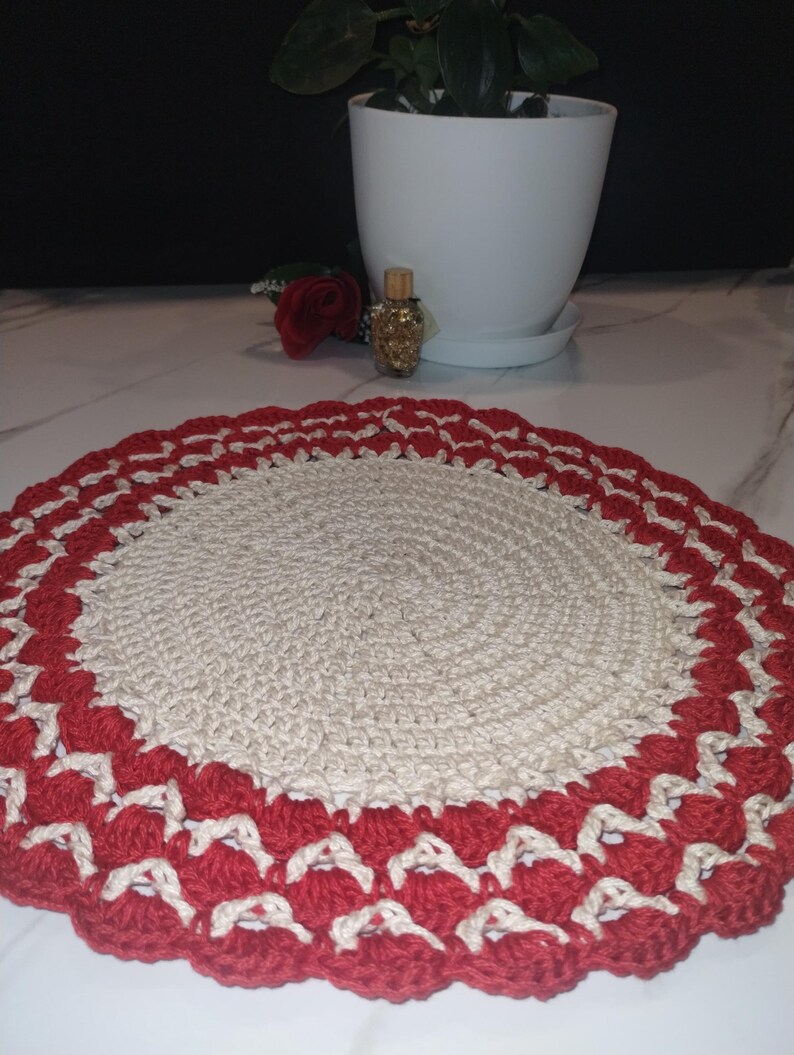 May include: A handmade, round crochet doily. The center is a natural beige color, surrounded by a scalloped border of red and cream-colored stitches. The doily is likely used for decorative purposes.