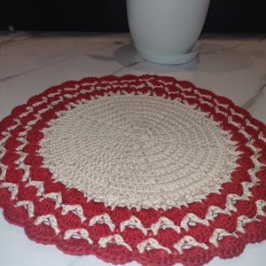 May include: A handmade, circular crochet doily. The center is a natural beige color, surrounded by a scalloped border of red and white. The doily is likely used as a decorative item or a coaster.