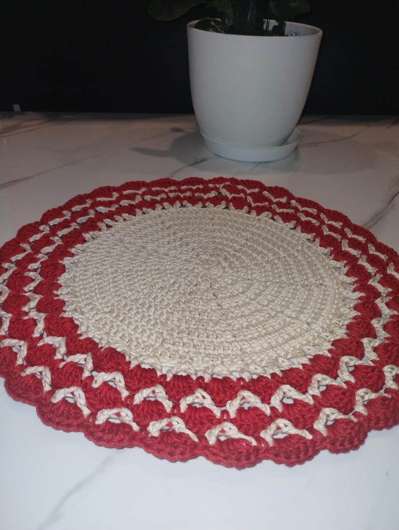 May include: A handmade, circular crochet doily. The center is a natural beige color, surrounded by a scalloped border of red and white. The doily is likely used as a decorative item or a coaster.