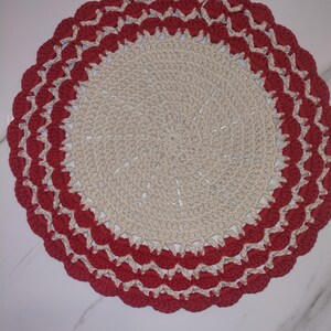May include: A handmade, circular crochet doily. The center is a light beige color, with a scalloped border in red and cream. The doily is likely used for decorative purposes.