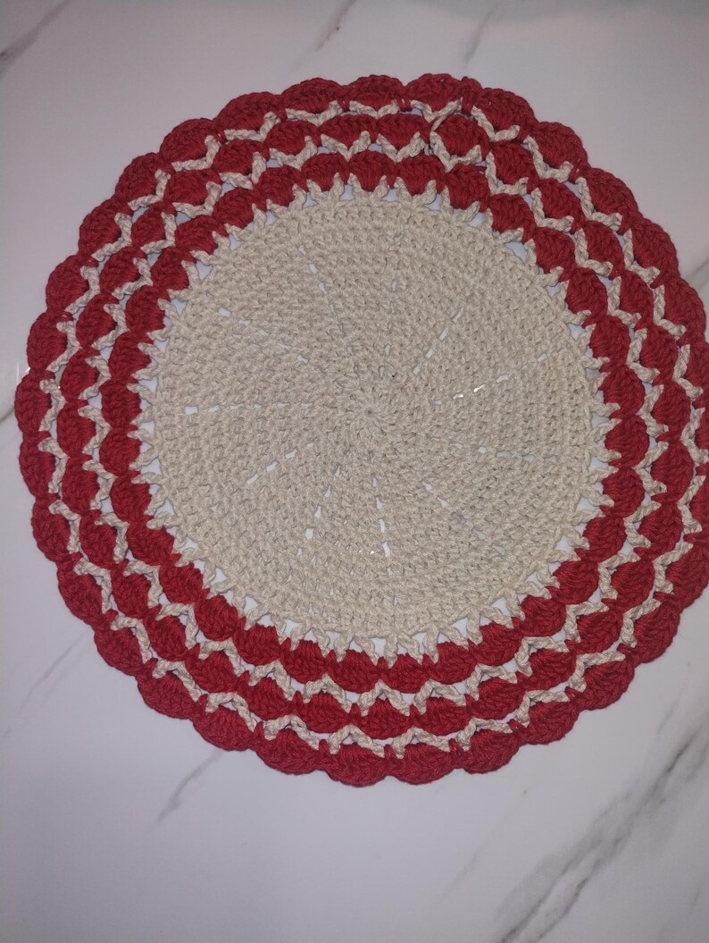 May include: A handmade, circular crochet doily. The center is a light beige color, with a scalloped border in red and cream. The doily is likely used for decorative purposes.