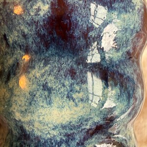 以下が含まれることがあります： ユニークなテクスチャード釉薬のセラミック花瓶。青、ティール、茶色の色が混ざり合い、マーブル模様を作り出しています。光沢のある表面は光を反射し、釉薬の複雑な模様を際立たせています。
