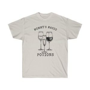May include: A light gray t-shirt with the text "Mommy's Magic Potions" printed in black. The text is surrounded by a drawing of three wine glasses, including a champagne flute.