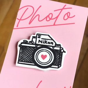 Könnte beinhalten: Eine rosa Karte mit den Worten "Photo glow" in Kursivschrift. Auf der Karte befindet sich eine Schwarzweißillustration einer Kamera mit einem roten Herzen im Objektiv. Die Kamera trägt die Aufschrift "Nikon".