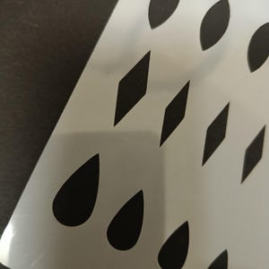 Peut inclure: Un pochoir en plastique blanc avec des formes découpées variées, notamment des gouttes, des losanges et des ovales. Le pochoir est incliné, révélant la surface sombre en dessous et mettant en valeur l'espace négatif.