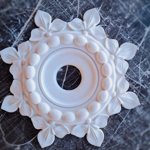May include: White plaster ceiling medallion with a circular design featuring a ring of raised ovals and a central hole. The medallion is decorated with eight stylized leaf shapes around the outer edge.