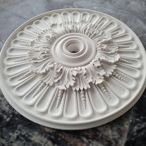 May include: White ceiling medallion with a decorative floral design and a central hole. The medallion is made of plaster and has a smooth, matte finish.