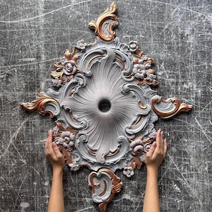 May include: A decorative ceiling medallion featuring a central circular opening surrounded by intricate gray scrollwork and floral relief patterns. The design is accented with metallic copper highlights on the raised edges and floral elements.