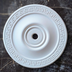 May include: A white ceiling medallion with a Greek key design around the outer edge. The center has a circular depression with a hole. The medallion is made of a smooth, matte material.