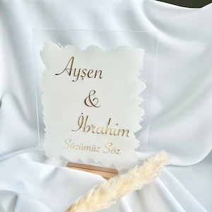 Könnte beinhalten: Ein klares Acrylschild mit goldenen Buchstaben, das die Hochzeit von Aysen & Ibrahim ankündigt. Das Schild lautet "Aysen & Ibrahim Sözümüz Söz", was übersetzt "Unser Wort ist unser Wort" bedeutet.