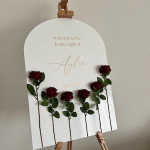 May include: An arched white acrylic welcome sign for a henna night featuring the text "welcome to the henna night of Aylin" and "May 03th, 2025" in gold lettering. Six dark red artificial roses are attached to the front of the sign.