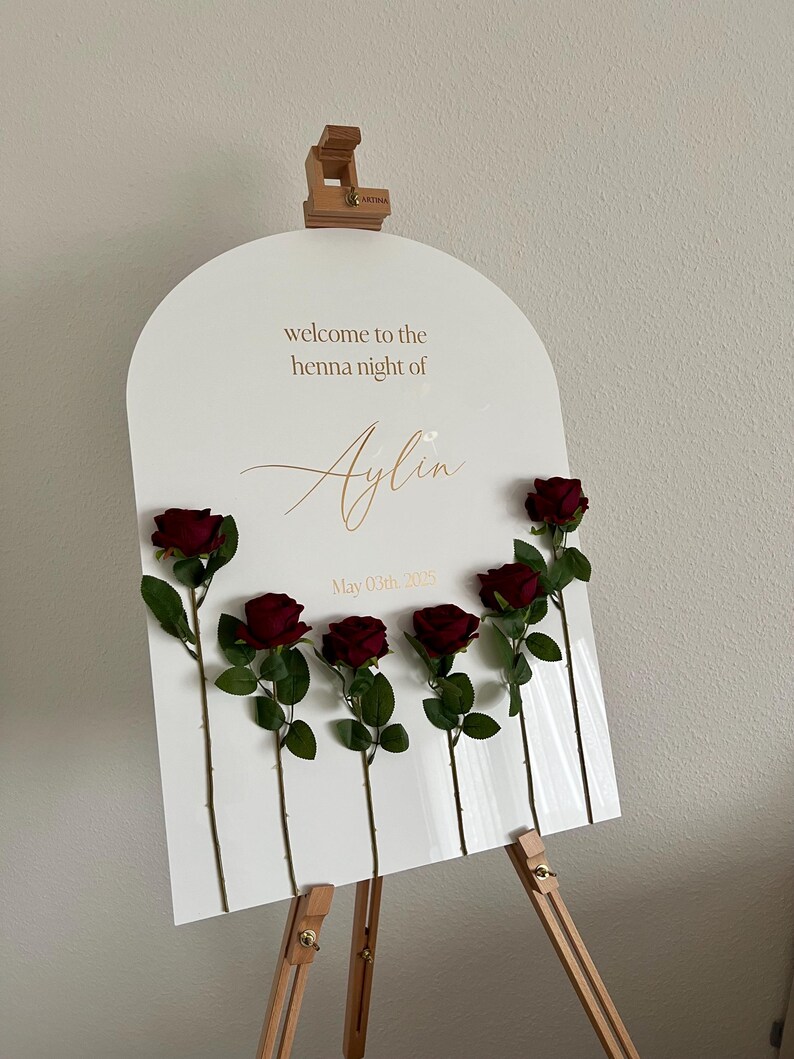 May include: An arched white acrylic welcome sign for a henna night featuring the text "welcome to the henna night of Aylin" and "May 03th, 2025" in gold lettering. Six dark red artificial roses are attached to the front of the sign.