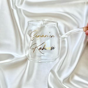 May include: A clear glass mug with a rounded body and a side handle. The front of the mug features gold cursive text that reads Sinan'in Kahvesi. The glass is transparent, showcasing a smooth, curved design with a small spout on the top rim.