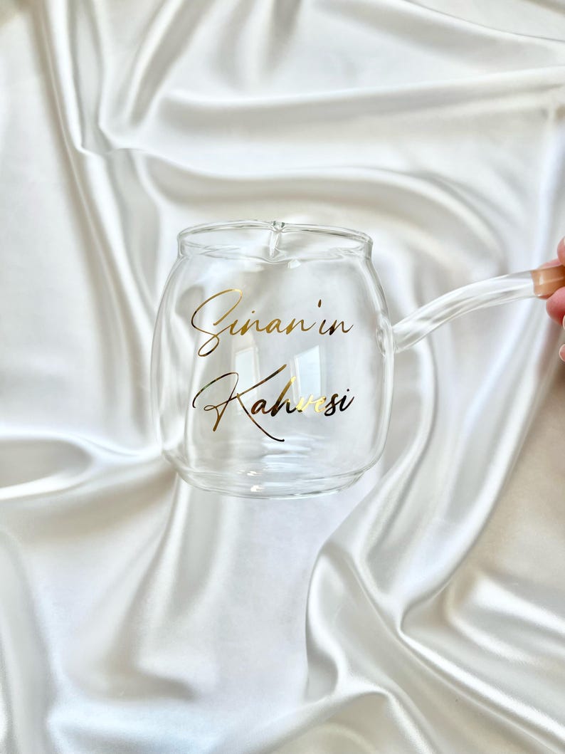 May include: A clear glass mug with a rounded body and a side handle. The front of the mug features gold cursive text that reads Sinan'in Kahvesi. The glass is transparent, showcasing a smooth, curved design with a small spout on the top rim.