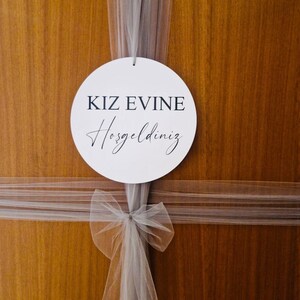 May include: A white circle sign with black text that reads "KIZ EVINE Hosgeldiniz" hanging on a wooden door with a white tulle bow.