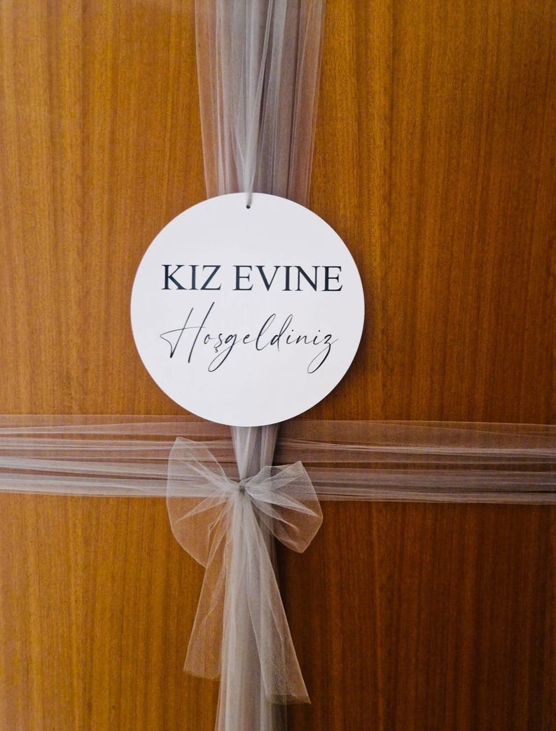 May include: A white circle sign with black text that reads "KIZ EVINE Hosgeldiniz" hanging on a wooden door with a white tulle bow.
