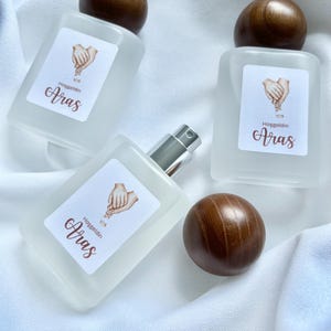 May include: Three frosted glass perfume bottles with wooden spherical caps. Each bottle has a white label with the word "Aras" and a graphic of two hands. The bottles are arranged on a white fabric surface.