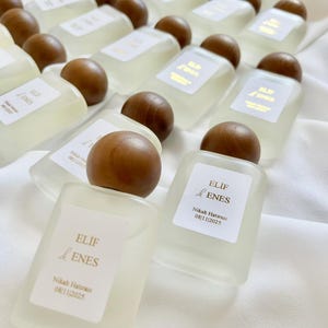 May include: A collection of small, rectangular perfume bottles with wooden caps. Each bottle has a white label with gold text that reads "ELIF & ENES" and the date "08/11/2025". The bottles are arranged on a white fabric surface.