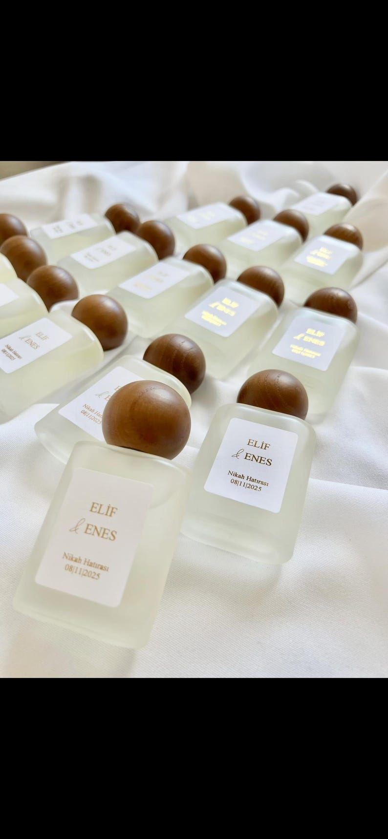May include: A collection of small, rectangular perfume bottles with wooden caps. Each bottle has a white label with gold text that reads "ELIF & ENES" and the date "08/11/2025". The bottles are arranged on a white fabric surface.