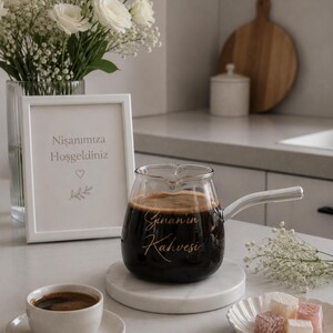 May include: A clear glass coffee pot with a silver handle, filled with dark coffee and featuring the gold script text "Sinan'ın Kahvesi" on the front. The pot sits on a round white marble coaster next to a small white cup and saucer.