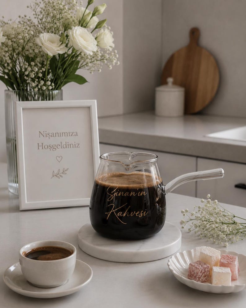 May include: A clear glass coffee pot with a silver handle, filled with dark coffee and featuring the gold script text "Sinan'ın Kahvesi" on the front. The pot sits on a round white marble coaster next to a small white cup and saucer.