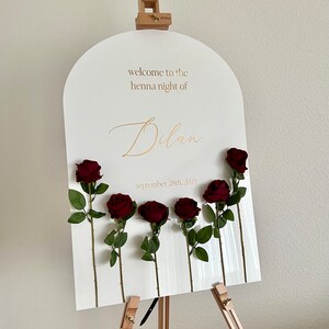 May include: A white arched acrylic welcome sign on a wooden easel featuring gold text that reads "welcome to the henna night of Dilan" and "september 28th, 2024". Six dark red artificial roses with green stems and leaves are attached to the sign.