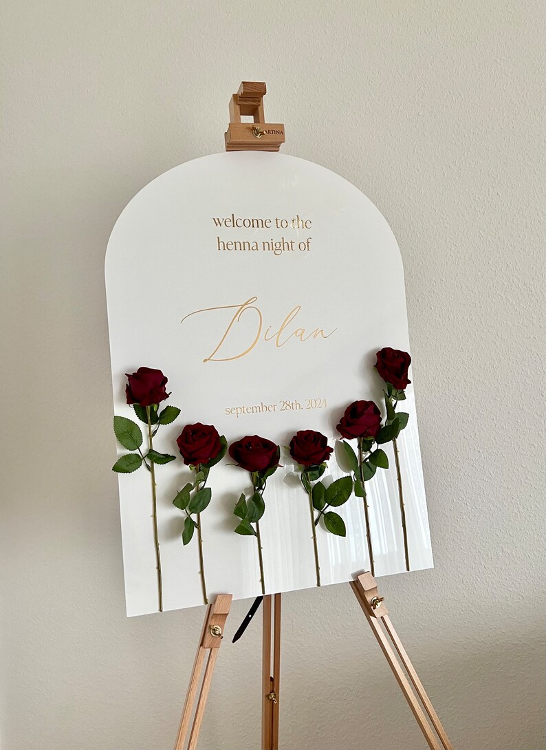 May include: A white arched acrylic welcome sign on a wooden easel featuring gold text that reads "welcome to the henna night of Dilan" and "september 28th, 2024". Six dark red artificial roses with green stems and leaves are attached to the sign.