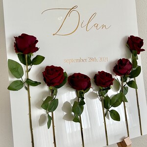 May include: An arched white sign featuring the gold text "welcome to the henna night of Dilan" and "september 28th, 2024". Six dark red artificial roses with green stems and leaves are attached to the front of the sign, arranged in a horizontal row.