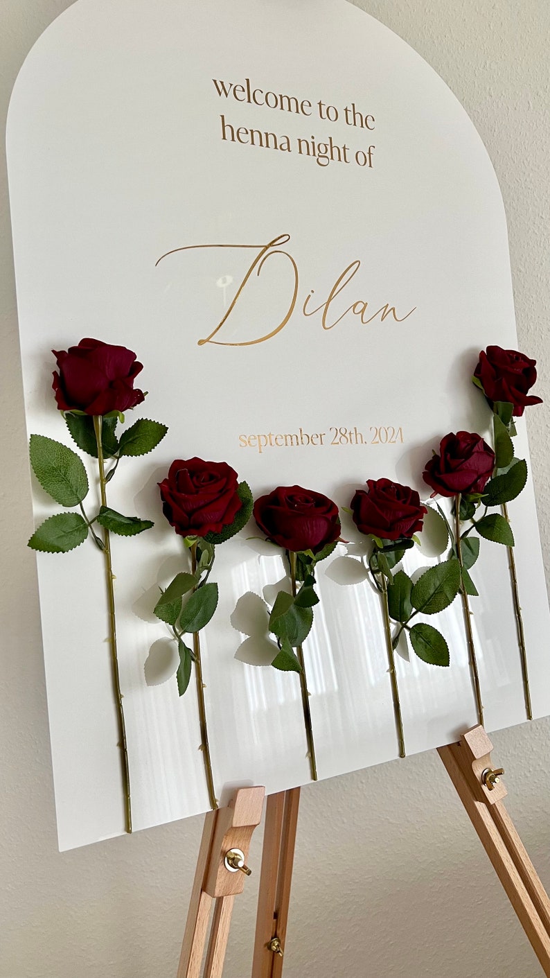 May include: An arched white sign featuring the gold text "welcome to the henna night of Dilan" and "september 28th, 2024". Six dark red artificial roses with green stems and leaves are attached to the front of the sign, arranged in a horizontal row.
