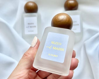 Puede incluir: Frascos de perfume de vidrio esmerilado con tapones de madera. Cada frasco tiene una etiqueta con los nombres "SEZEN y MAHIR" y la fecha "10.10.2025" en letras doradas. Los frascos están dispuestos sobre un fondo de tela blanca.