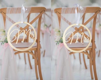 May include: Two wooden chairs with white tulle and pink ribbon decorations. A gold circle sign with the words "Mr." and "Mrs." hangs on each chair.