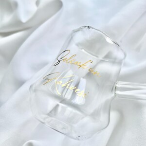 May include: A clear glass mug with a double-walled design and a handle, featuring gold cursive text that reads "Gelenk'in Kahvesi" printed on the front surface. The mug has a smooth, rounded shape and a transparent appearance.
