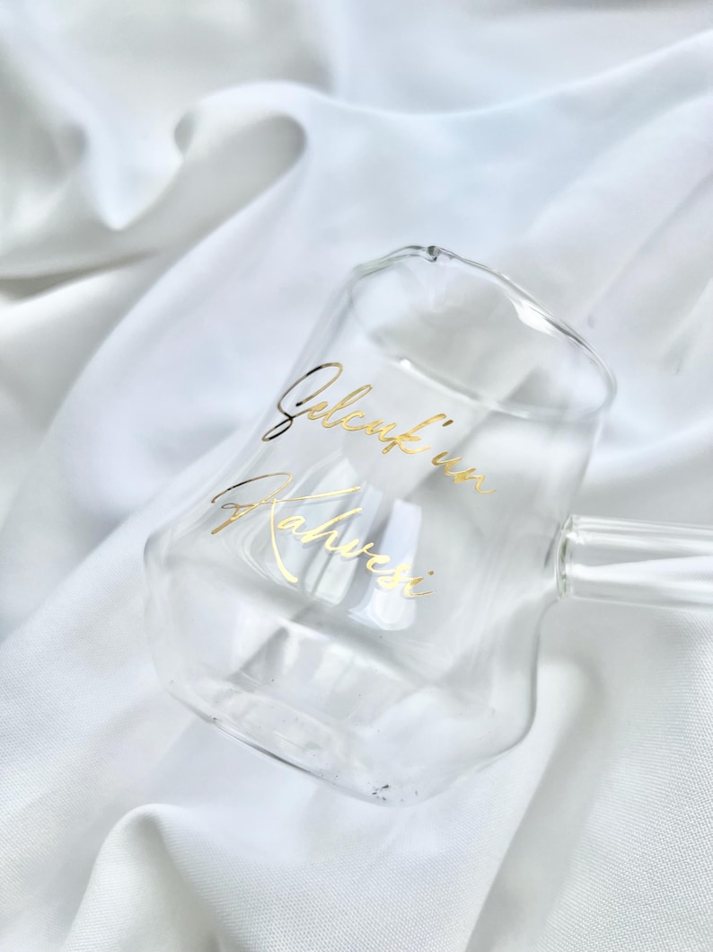May include: A clear glass mug with a double-walled design and a handle, featuring gold cursive text that reads "Gelenk'in Kahvesi" printed on the front surface. The mug has a smooth, rounded shape and a transparent appearance.