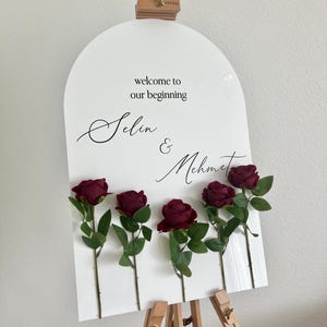 May include: An arched white acrylic sign featuring the black text "welcome to our beginning" and the names "Selin & Mehmet" in a script font. Five dark red artificial roses with green stems and leaves are attached to the front of the sign.