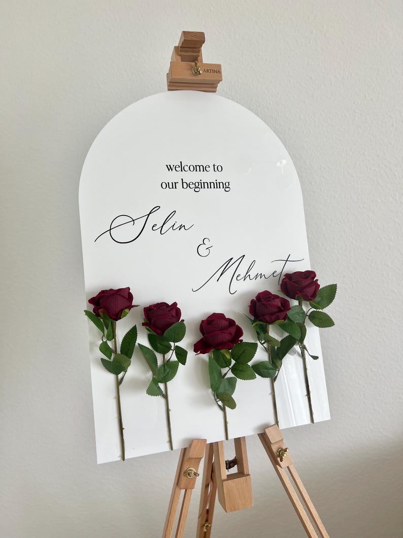May include: An arched white acrylic sign featuring the black text "welcome to our beginning" and the names "Selin & Mehmet" in a script font. Five dark red artificial roses with green stems and leaves are attached to the front of the sign.
