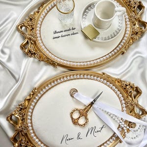 Peut inclure: Deux plateaux ovales dorés avec des centres en velours blanc. L'un contient un verre et une tasse à thé avec un accent doré, l'autre des ciseaux dorés et un ruban blanc. Les deux plateaux ont des bordures de perles et des accents floraux. Du texte est visible.