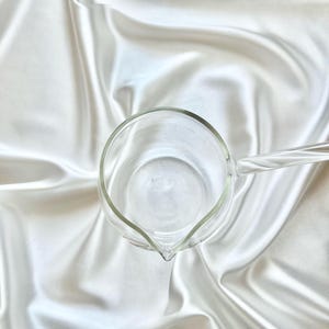 May include: A clear glass pitcher with a round body and a single handle extending from the side. The pitcher features a small, pointed spout for pouring and a smooth, transparent surface, viewed from a top-down perspective.