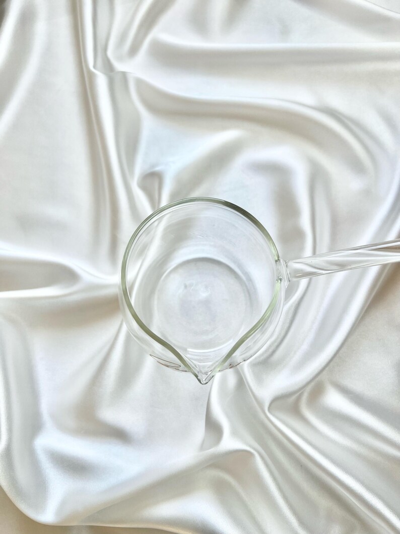 May include: A clear glass pitcher with a round body and a single handle extending from the side. The pitcher features a small, pointed spout for pouring and a smooth, transparent surface, viewed from a top-down perspective.