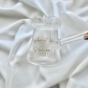 May include: A clear glass coffee pot with a ribbed texture and a long handle. The front of the pot features gold cursive text that reads Ahmet in Kahvesi. The vessel has a wide base, a narrow neck, and a small pouring spout at the top rim.