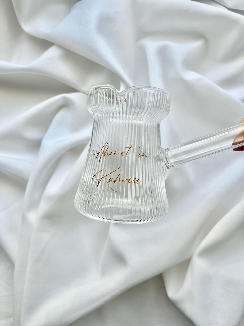 May include: A clear glass coffee pot with a ribbed texture and a long handle. The front of the pot features gold cursive text that reads Ahmet in Kahvesi. The vessel has a wide base, a narrow neck, and a small pouring spout at the top rim.