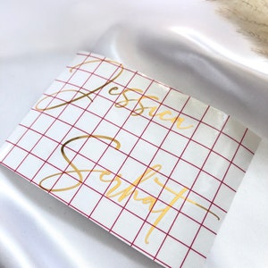 May include: A white sheet of paper with a red grid pattern. The names "Jessica" and "Schmat" are written in gold cursive lettering on the paper.