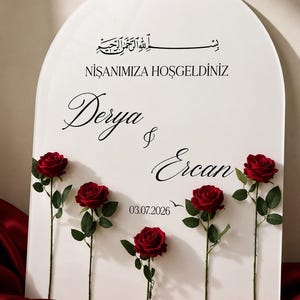 May include: An arched white sign featuring black text that reads "NİŞANIMIZA HOŞGELDİNİZ", "Derya & Ercan", and "03.07.2026" with Arabic calligraphy at the top. Five deep red roses with green stems and leaves are attached to the front of the sign.