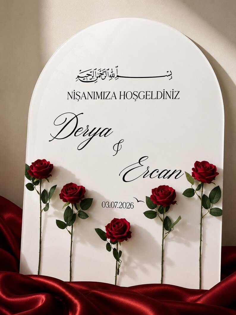 May include: An arched white sign featuring black text that reads "NİŞANIMIZA HOŞGELDİNİZ", "Derya & Ercan", and "03.07.2026" with Arabic calligraphy at the top. Five deep red roses with green stems and leaves are attached to the front of the sign.