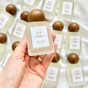 May include: A collection of small, frosted glass bottles with wooden caps. Each bottle has a label with gold text that reads "ELIF & ENES" and "Nikah Hatırası 08/11/2025". The bottles are arranged on a white surface.
