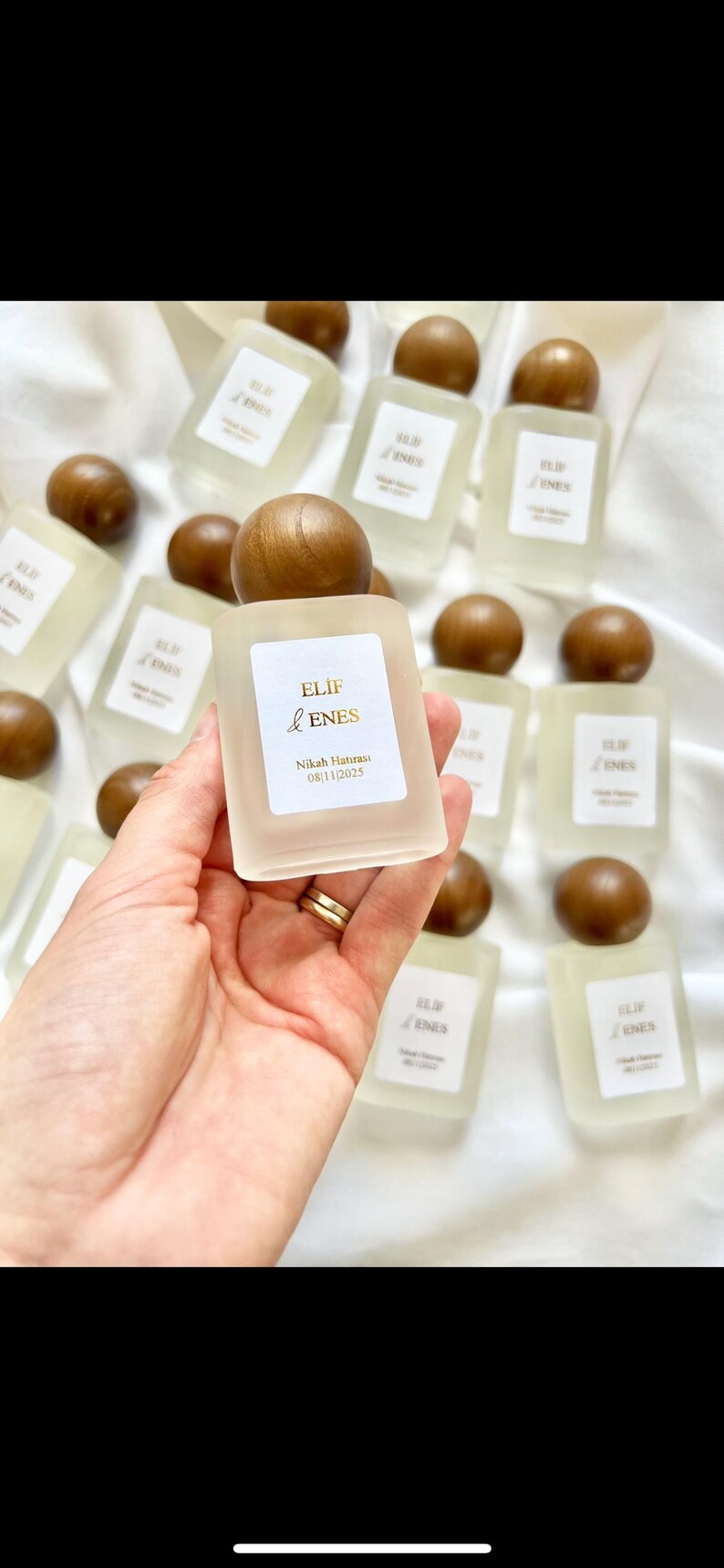 May include: A collection of small, frosted glass bottles with wooden caps. Each bottle has a label with gold text that reads "ELIF & ENES" and "Nikah Hatırası 08/11/2025". The bottles are arranged on a white surface.
