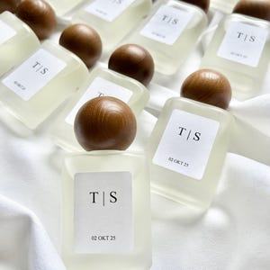 May include: A collection of small, rectangular perfume bottles with wooden spherical caps. Each bottle has a white label with the initials "T|S" and the date "02 OKT 25". The bottles are arranged on a white, textured surface.