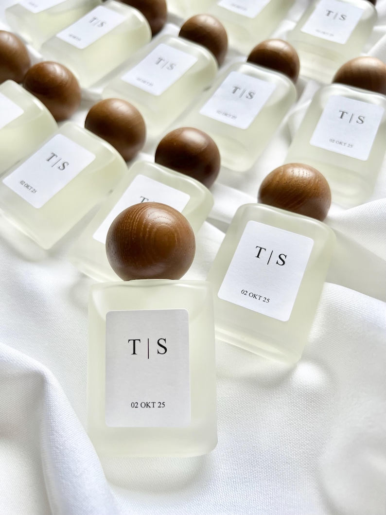 May include: A collection of small, rectangular perfume bottles with wooden spherical caps. Each bottle has a white label with the initials "T|S" and the date "02 OKT 25". The bottles are arranged on a white, textured surface.