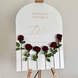 May include: An arched white acrylic welcome sign featuring the gold text "welcome to the henna night of Dilan" and "september 28th, 2024". Six dark red artificial roses with green stems and leaves are attached to the front of the sign.