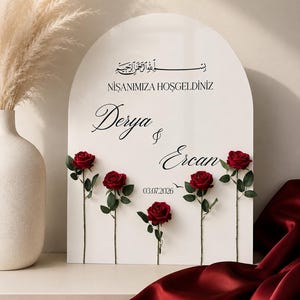 May include: An arched white sign featuring black text that reads "NİŞANIMIZA HOŞGELDİNİZ", "Derya & Ercan", and "03.07.2026" with Arabic calligraphy at the top. Five individual red roses with green stems and leaves are attached to the front surface.