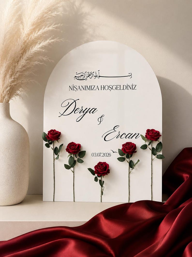 May include: An arched white sign featuring black text that reads "NİŞANIMIZA HOŞGELDİNİZ", "Derya & Ercan", and "03.07.2026" with Arabic calligraphy at the top. Five individual red roses with green stems and leaves are attached to the front surface.
